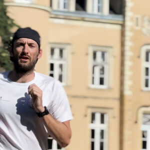 A man in a white shirt and black headband is jogging outdoors in front of a beige building with multiple white-framed windows. The background is slightly out of focus.