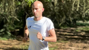 A man with a shaved head, wearing a white t-shirt and black watch, jogs outdoors on a sunny day. Trees and dappled sunlight are visible in the background.