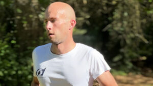 A man with a shaved head, wearing a white t-shirt, is running outdoors in a sunlit, wooded area. He appears focused and is sweating, suggesting exertion during exercise.