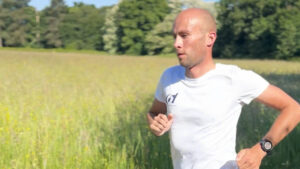 A man wearing a white shirt and a black watch is running outdoors through a grassy field with trees in the background on a sunny day.