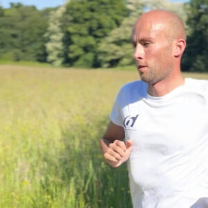A man wearing a white shirt and a black watch is running outdoors through a grassy field with trees in the background on a sunny day.