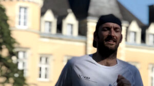 A man in a white shirt and backwards cap runs outdoors in front of a blurred yellow building with pointed rooftops and windows.