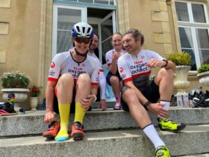 Four cyclists in matching “Team Oxygen Addict” jerseys sit and smile on stone steps outside a building, with helmets, cycling shoes, and water bottles visible around them. Two people sit behind, making peace signs and smiling.