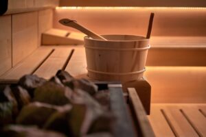 A wooden sauna room with a bench, a bucket, a ladle, and hot stones, softly lit by warm ambient lighting.