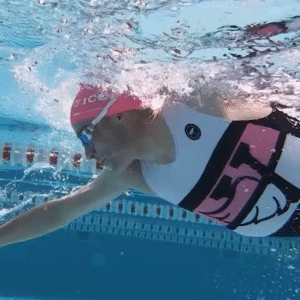 Underwater view of a swimmer in a pink swim cap and patterned swimsuit, reaching forward with one arm while swimming freestyle in a clear blue pool near a lane divider.