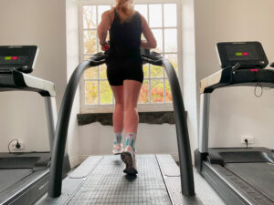 A person with long hair in a ponytail runs on a treadmill in a bright room, facing a large window with trees visible outside. Two other treadmills are on either side. The runner wears black workout clothes and colorful socks.