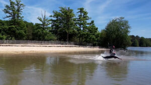 A person wearing a helmet rides a jet ski quickly across a calm, shallow lake near a sandy shore lined with trees under a bright blue sky.