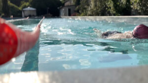 A person swimming in an outdoor pool is seen from water level; another hand in the foreground holds a floating object, pointing toward the swimmer. Trees and buildings are visible in the background.