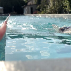 A person swimming in an outdoor pool is seen from water level; another hand in the foreground holds a floating object, pointing toward the swimmer. Trees and buildings are visible in the background.
