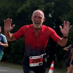 An older man wearing a red shirt with race number 295 smiles joyfully and raises both hands while running in a race. Other runners and a traffic cone are visible in the background.