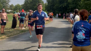 A smiling runner gives two thumbs up while participating in a race, wearing a blue jersey and bib number 296. Other racers and spectators line the path, cheering on the athletes under a sunny sky.