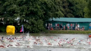 A group of swimmers wearing swim caps compete in an open water race, creating splashes in a river. Spectators watch from the grassy, tree-lined shore in the background. A yellow buoy and pink flag are visible near the swimmers.