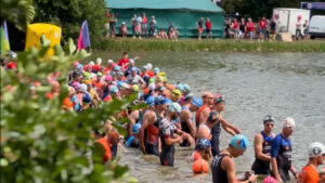 A large group of swimmers wearing colorful swim caps and wetsuits waits at the edge of a lake for a race to begin, with spectators watching from the shore and a green tent in the background.