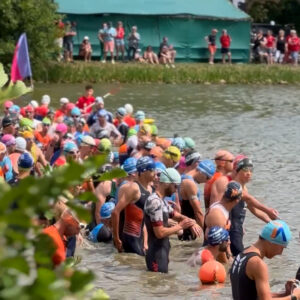 A large group of swimmers wearing colorful swim caps and wetsuits waits at the edge of a lake for a race to begin, with spectators watching from the shore and a green tent in the background.