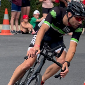 A cyclist wearing a helmet and sunglasses makes a sharp turn on a road during a race, with orange traffic cones and spectators watching from the side.