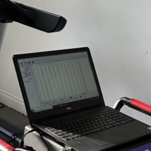 A Dell laptop displaying a data spreadsheet is placed on the control panel of a treadmill, with sunlight illuminating part of the scene. The treadmill’s armrests and a portion of the console are also visible.