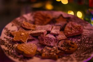 A decorative plate filled with assorted holiday cookies, including star and heart shapes, dusted with powdered sugar. Warm, festive lights blur in the background, creating a cozy atmosphere.