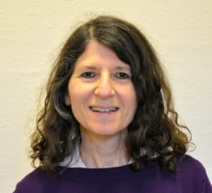 A woman with wavy brown hair and a friendly smile is wearing a purple sweater over a collared shirt, standing in front of a plain light-colored wall.