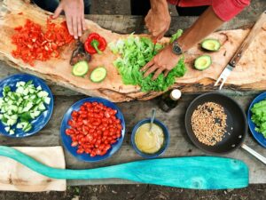 Two people prepare fresh vegetables, including lettuce, avocado, tomatoes, cucumber, and red pepper, on a rustic wooden board, surrounded by bowls of ingredients and a pan with corn on a wooden table outdoors.
