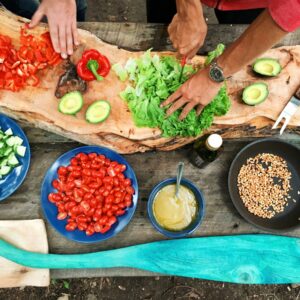 Two people prepare fresh vegetables, including lettuce, avocado, tomatoes, cucumber, and red pepper, on a rustic wooden board, surrounded by bowls of ingredients and a pan with corn on a wooden table outdoors.