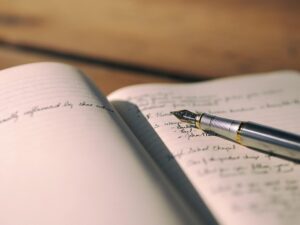 A close-up of an open notebook with handwritten notes and a silver fountain pen resting on the pages. The background is softly blurred, suggesting a calm and focused writing environment.