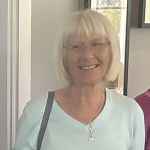 Smiling older woman with short white hair and glasses, wearing a light blue top and a silver necklace, standing indoors near a doorway.