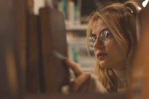 A young woman with glasses and long blonde hair looks closely at books on a shelf in a library, reaching out to select one.