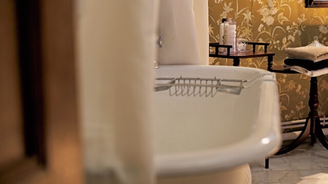 Close-up of a vintage-style bathroom with a white clawfoot bathtub. The wall features floral-patterned wallpaper. A small table beside the tub holds bath supplies, and a white shower curtain is partially visible.