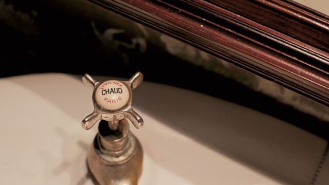 Close-up of a vintage faucet handle labeled CHAUFFÉ CHAUD PARIS in a bathroom setting, featuring a cross-shaped design. The faucet is attached to a white sink with a dark wooden trim visible at the top.