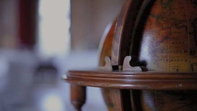A close-up of a vintage globe partially open in a dimly lit room. The wooden frame is in focus, with blurred background suggesting a cozy interior space. The globes surface displays parts of continents and old-style text.