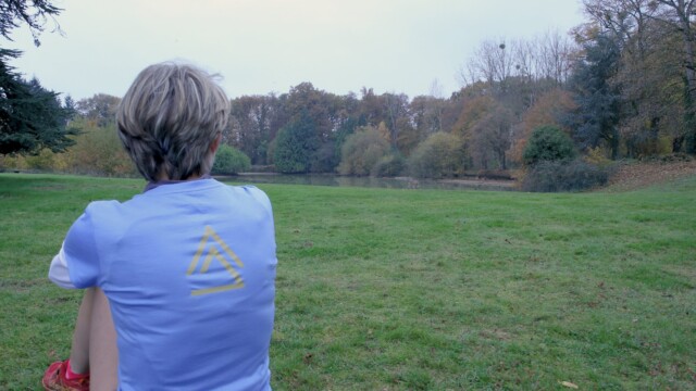 Sitting on a mat in a park, someone with short hair and a blue shirt featuring a yellow triangle design enjoys the view of a grassy field and tree-lined pond under an overcast sky, reminiscent of contemplating during 1-1 Sessions.
