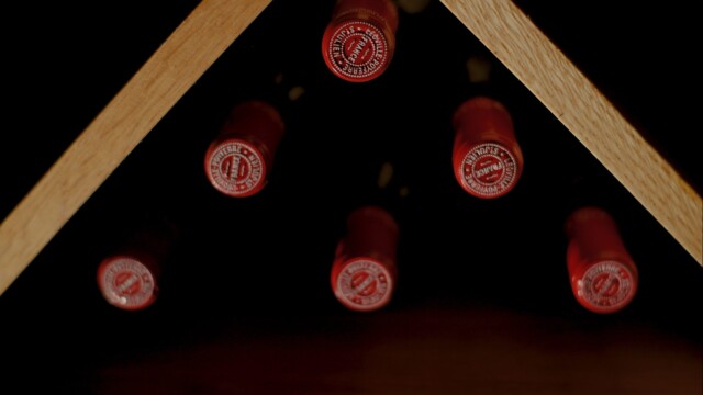 Bottles of Club Socials red wine are horizontally stored in a triangular wooden rack, showcasing red caps with white text. The neatly aligned caps create a visually appealing display.