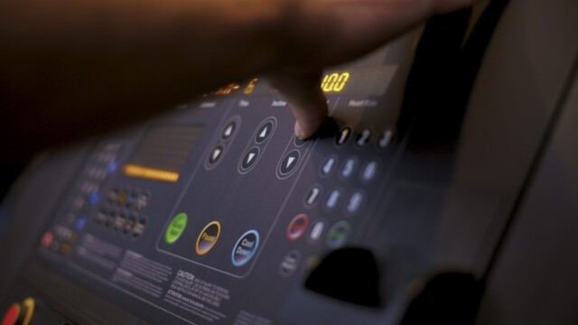 A person’s hand pressing buttons on a treadmill console with various controls and digital displays in a dimly lit setting.