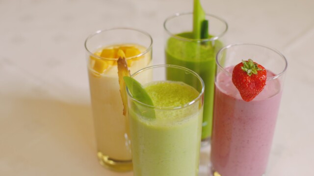 Four smoothies in glasses on a light surface: a yellow mango smoothie with a slice, a green smoothie garnished with greens, a darker green veggie smoothie, and a pink strawberry smoothie topped with fruit.