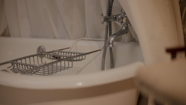 A white freestanding bathtub with a silver clawfoot faucet is partially covered by a white curtain. A metal rack rests across the tubs edges, and a flexible shower hose hangs from the faucet. The scene suggests a cozy, vintage bathroom setting.