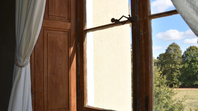 A sunlit wooden-framed window with white curtains is partially open, revealing a view of lush green trees and a blue sky with scattered clouds outside. The wall beside the window is cream-colored.