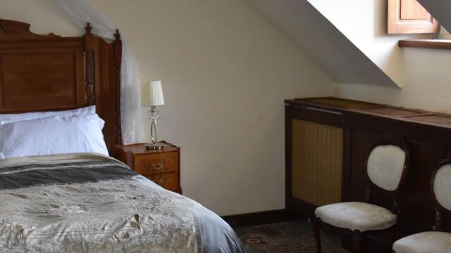 A cozy attic bedroom with a sloped ceiling featuring a wooden bed with white and gray bedding. Theres a small nightstand with a lamp beside the bed, and two elegant chairs are placed against the wall near a wooden cabinet. A window offers natural light.