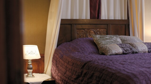 A cozy bedroom with a dark wooden headboard and a purple quilted bedspread. Three patterned pillows are neatly arranged on the bed. A decorative lamp sits on a bedside table, casting a warm glow. Cream-colored drapes hang in the background.
