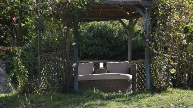 A cozy outdoor gazebo with a round wicker couch sits in a lush garden. Pink roses climb the wooden frame, and greenery surrounds the shelter, creating a peaceful, inviting atmosphere.