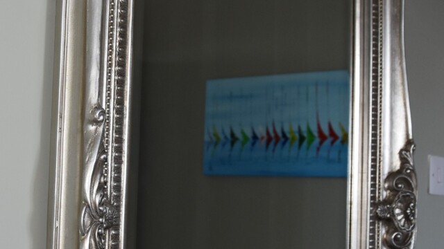 A large, ornate silver mirror reflects a section of a room with a painting on the wall. The painting features abstract colorful arrows on a blue background. A radiator can be seen below the painting. The rooms walls are light green.