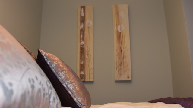 The art of the matter Close-up of two decorative pillows on a bed with a white comforter. The wall behind is painted in a soft gray color and features two vertical floral paintings with brown and beige tones. The room has a calm and minimalist aesthetic.