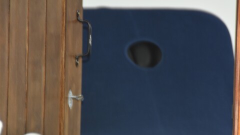 A partially open wooden door with visible hinges and a metal handle. Through the doorway, a dark blue chair with an oval cutout in the backrest is partially visible against a white background.