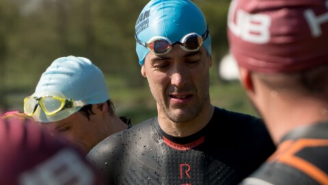 A group of swimmers in wetsuits are gathered outdoors. One central swimmer wears a blue swim cap and goggles. Others wear red caps. The background is blurred, hinting at trees and an outdoor setting.