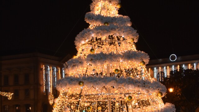 A festive Christmas tree adorned with glowing white lights and golden baubles stands illuminated at night. The tree is cone-shaped, decorated with fluffy white layers, and topped with a lit ring, set against a backdrop of dimly visible buildings.