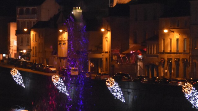 Night view of a riverside city with illuminated buildings. A large neon sign reads LAVAL atop a building. Festive lights decorate the riverside, reflecting in the water. Water fountains are lit with colorful lights.