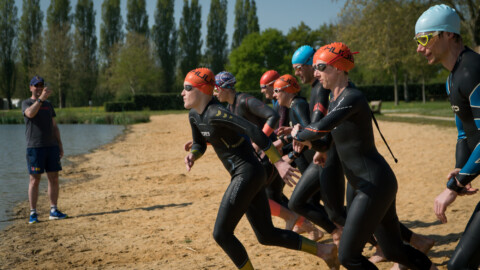A group of triathletes in wetsuits and colorful swim caps run across a sandy beach towards the water at our local lake, preparing to start a pratcice race during our festival of endurance. A person on the left, wearing a cap and shorts, appears to give instructions or start the race. Trees and grass are in the background.