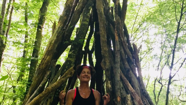 A person sits cross-legged and meditating inside a tipi-like structure made of sticks in a lush green forest. They wear a black tank top and shorts, with eyes closed and hands in a relaxed yoga pose. Sunlight filters through the trees.