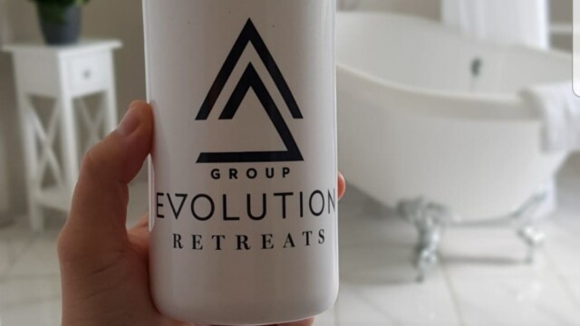 A hand holds a white Group Evolution Retreats water bottle with a black lid. In the background, theres a white, clawfoot bathtub, white curtains, and a small table with a potted plant, all set on a white tiled floor.