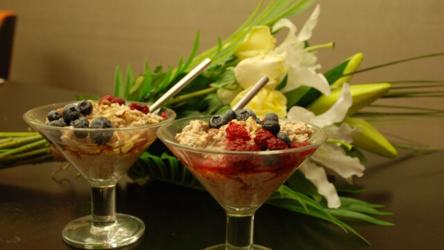 Two dessert glasses filled with a mixture of oats, yogurt, and fresh berries, including blueberries and raspberries, are placed on a dark table. Elegant white lilies and green foliage lie in the background.