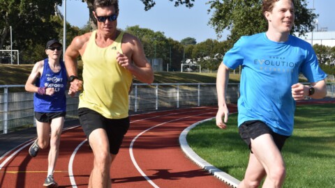 Three people running on our local athletics track during the run well retreat. One wears a yellow tank top, another a blue shirt with Group Evolution Retreats emblazoned on the front, and the third a blue tank top with a hat. Trees and a railing line the track. Its a very sunny day.
